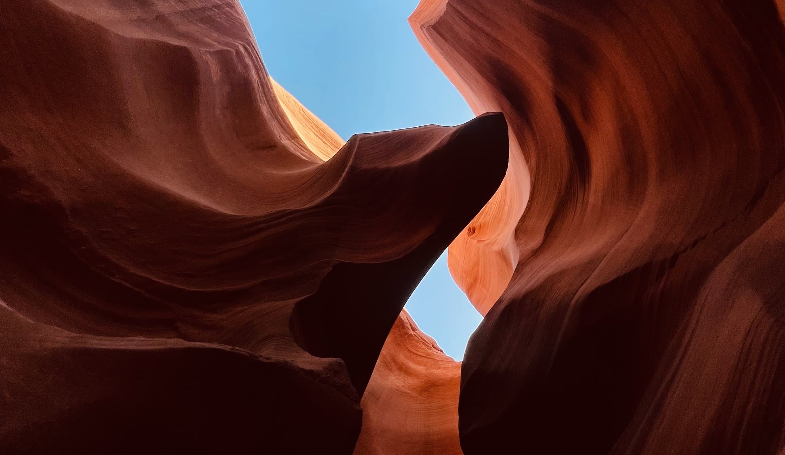 มองขึ้นไปเห็นท้องฟ้าสีฟ้าผ่านผนังหินทรายสีส้มโค้งเป็นคลื่นใน Antelope Canyon รัฐแอริโซนา